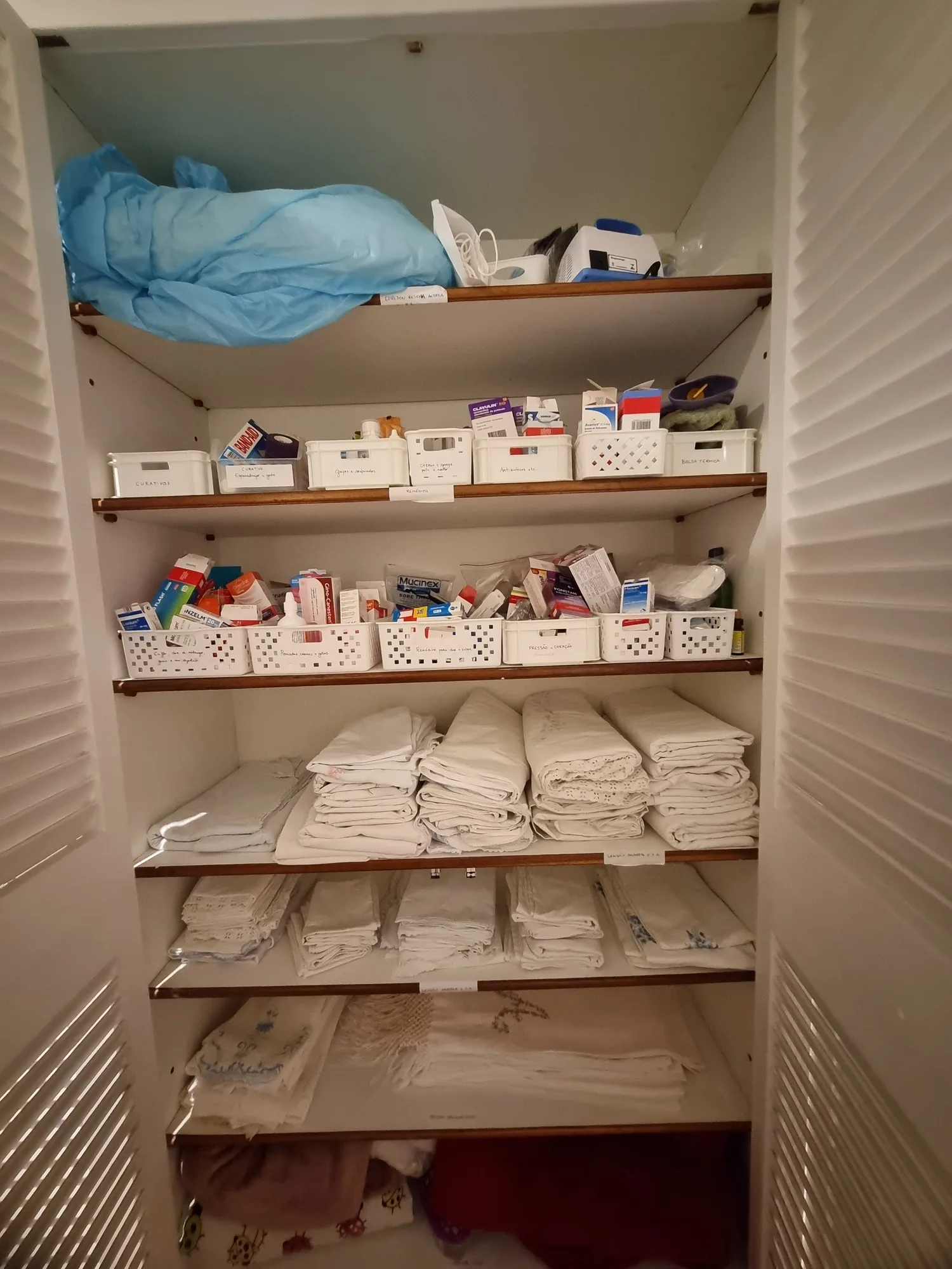 Armário de corredor antes da organização, com medicamentos soltos nos cestos e roupa de cama dobrada sem separação